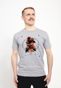Camiseta de algodón gris con un diseño gráfico floral en rojo y verde, centrado en el pecho con elementos de texto en negro. Mangas cortas.