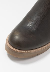 Tmavě hnědá kožená kotníková bota s kulatou špičkou, strukturovaným povrchem a béžovým šitím. Má pevnou světle hnědou gumovou podrážku.