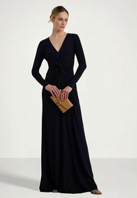 Γυναίκα με μαύρο μακρύ φόρεμα με στροβιλισμένο μπροστινό σχέδιο, κρατώντας μια τακτική τσάντα, στέκεται απέναντι σε ένα απλό ανοιχτό φόντο.