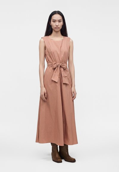 Femme aux longs cheveux noirs porte une robe midi sans manches rose poudré avec ceinture nouée en nœud et bottines marron, debout devant un fond uni.