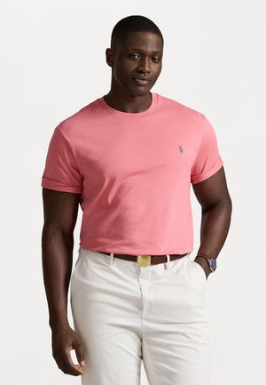 Camiseta de algodón de manga corta en color coral, con cuello redondo y un pequeño logo azul en el lado izquierdo del pecho. Combinada con pantalones blancos.