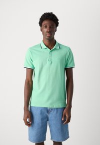 Camiseta polo verde menta con tapeta de dos botones, cuello a rayas y bordes de las mangas a rayas, combinada con pantalones cortos de mezclilla azul claro. Sin patrones visibles.