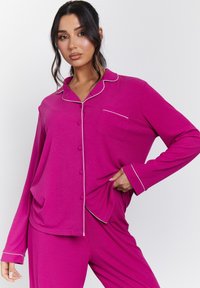 Conjunto de pijama de dos piezas en color rosa con mangas largas, cierre de botones en la parte delantera y ribetes blancos en el cuello y los puños; cuenta con un bolsillo en el pecho.