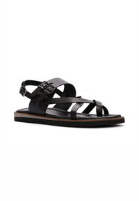 Sandal de cuero negro con diseño de tiras entrecruzadas, hebillas ajustables y una suela plana con una banda de acento marrón. Textura suave.