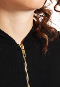 Musta huppari, jossa on kullanvärinen vetoketju kaulassa, valmistettu pehmeästä kankaasta, jolla on sileä pinta ja istuva muotoilu kauluksen ympärillä.