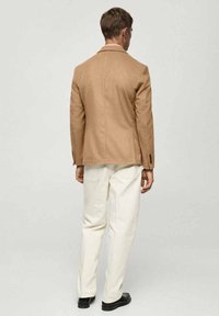 Bež vuneni sako s prilagođenim krojem, zatvaranjem na dva gumba i džepovima s preklopom, uparen s bijelim hlačama i crnim cipelama.