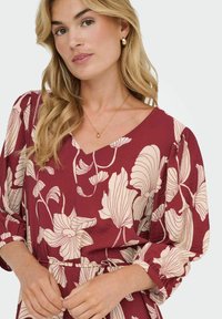 Burgundiklänning med blommig beige tryck, v-ringning och puffärmar; modellen accessoriserade med guldörhängen och ett fint guldhalsband.