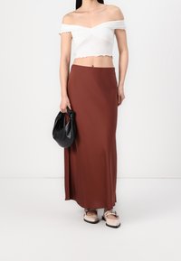 Hnědá, těsně přiléhající maxi sukně s hladkým povrchem, v kombinaci s bílým topem s odhalenými rameny. Drží černou uzlovou kabelku a nosí světlou obuv.