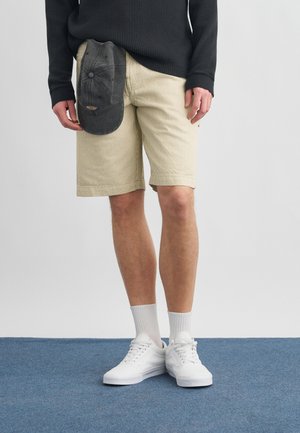 Muž v béžových manšestrových šortkách, bielych teniskách, bielych ponožkách a čiernej dlhorukávovej košeli drží tmavosivú baseballovú čiapku po boku.