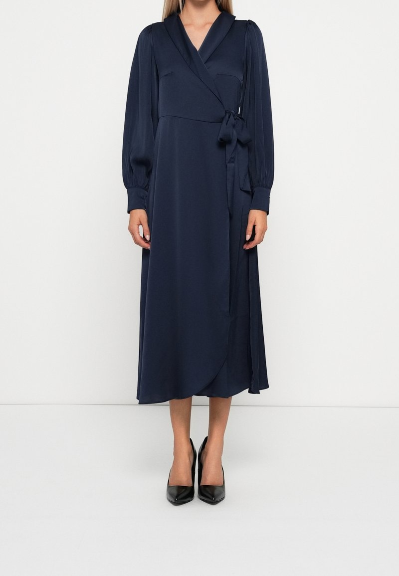 Femme portant une robe portefeuille bleu marine à manches longues avec une ceinture sur le côté et des escarpins noirs à bout pointu, debout devant un fond blanc uni.