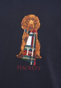 Rjav pes, ki nosi večbarvni karirasti šal z izvezeno črko "H" nad besedo "Hackett" na temnem materialu.