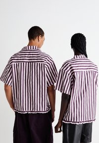 Camisa de mangas curtas em riscas verticais roxas e brancas, com colarinho pontudo e bainha com babados. O tecido parece leve e suave.