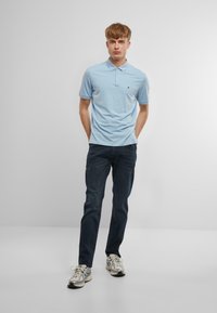 Gaiši zilas polo krekls ar īsām piedurknēm un apkakli, apvienots ar tumši zilām džinsām un gaišiem sporta apaviem. Minimāla zīmola redzamība.