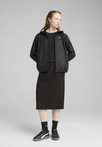 Veste légère noire avec capuche, portée par-dessus une robe sombre. Présente une fermeture éclair et le logo Puma. Assortie avec des baskets noires et des chaussettes.
