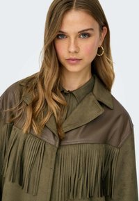 Giacca verde oliva con dettagli frangiati e inserti in pelle. Presenta un collo a camicia e una texture morbida, adatta per un abbigliamento casual.
