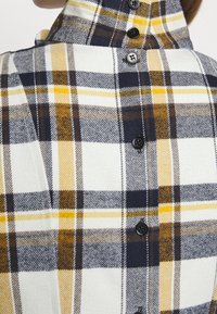 Long-sleeve checkered shirt in white, yellow, navy, and brown. Features a high collar and black buttons along the back. Soft, textured fabric.