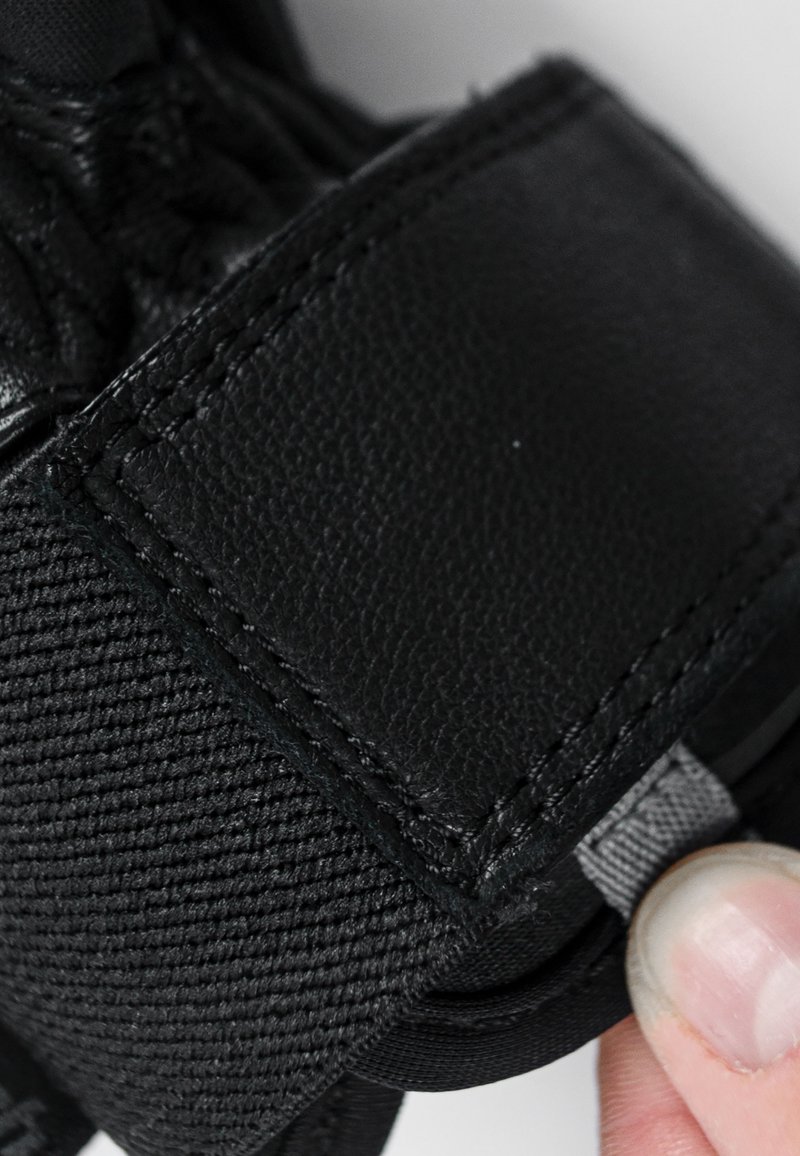 Schwarze Handschuhe aus strukturiertem Stoff mit einem Lederbesatz. Der Besatz hat sichtbare Nähte und eine graue Zuglasche zur Einstellung.