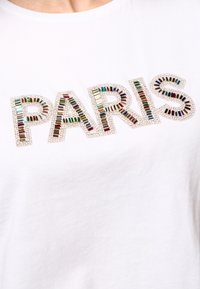 Witte katoenen t-shirt met het woord "PARIS" in kleurrijke sequins en een glinsterende rand, wat zorgt voor textuurcontrast op de gladde stof.