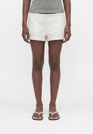 Άτομο που φοράει λευκά σορτς με γυρισμένο τελείωμα, γκρι αμάνικη μπλούζα και μπεζ σαγιονάρες, στέκεται μπροστά σε ένα απλό λευκό φόντο.