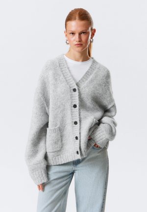 Jeune femme aux cheveux roux portant un cardigan à boutons gris clair, une chemise blanche, un jean bleu et des boucles d'oreilles créoles, debout avec une main dans la poche.