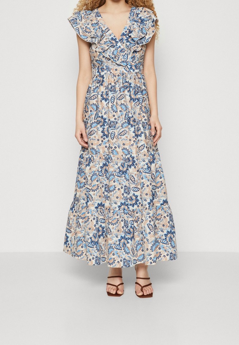 Robe maxi à motif floral bleu et beige, avec des manches volantées, un décolleté en V et une jupe à volants. Sandales brunes à lanières.