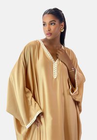 Beige kaftan med brede ærmer, V-halsudskæring og dekorativt hvidt kantbånd langs kanterne. Glat satin tekstur, flydende design.