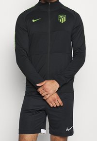 Chaqueta negra con cremallera y cuello alto, con detalles verdes y un logo. Combinada con pantalones cortos negros con ribete blanco y un logo en la pierna.