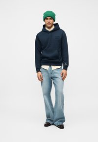 Homme portant un bonnet vert, un sweat à capuche bleu marine sur une chemise beige, un jean large bleu clair et des chaussures marron foncé, debout sur un fond blanc.