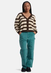 Gestreepte cardigan in bruin, crème en geel met een V-hals en knoopsluiting; gecombineerd met teal wijde broek en zwarte schoenen.