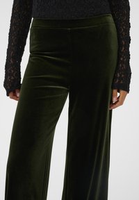 Pantaloni evazați din catifea verde, cu o textură netedă. Prezintă un talie înaltă și buzunare laterale. Asortați cu un top lung cu mâneci din dantelă neagră.