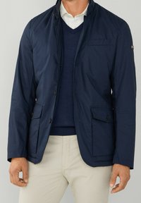 Marinblå lätt jacka med ståkrage, två framfickor och subtil textur. Bärs över en mörk tröja och ljusa byxor.