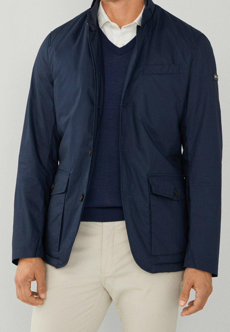 Marinblå lätt jacka med ståkrage, två framfickor och subtil textur. Bärs över en mörk tröja och ljusa byxor.