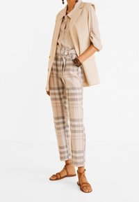 Beige bezrukávový blazer přes světle barevnou košili, doplněný kostkovanými kalhotami v béžové a hnědé. Hnědé sandály na pásky dotvářejí celý vzhled.