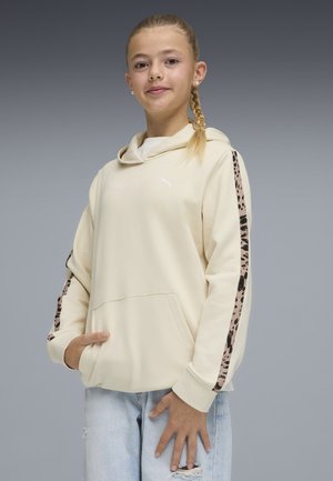 Mädchen mit geflochtenen Haaren, das einen cremefarbenen Kapuzenpullover mit gemusterten Streifen an den Ärmeln und hellblaue, zerrissene Jeans trägt, die Hände in den Taschen und auf dem Oberschenkel ruhend.