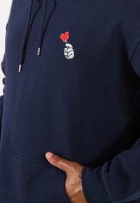 Persona che indossa un hoodie blu navy con un ricamo di una mano bianca che tiene un cuore rosso sul petto, con la mano che riposa nella tasca anteriore.