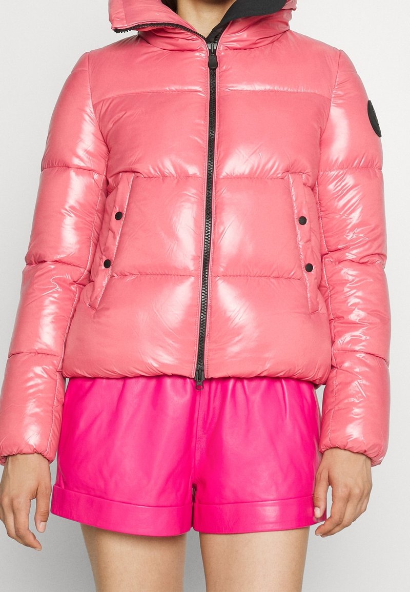 Chaqueta acolchada de color rosa brillante con una superficie brillante, cierre de cremallera y dos bolsillos frontales. Presenta una capucha y detalles en negro en los hombros.