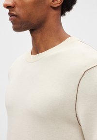 Gaišs bēša krāsas džemperis ar apaļu apkakli, ar gludu tekstūru un kontrastējošām brūnām šuvēm plecu šuvju līnijā.