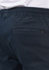 Marinblå byxor med en texturerad väv, som har ett elastiskt midjeband, bakficka och en liten logotyp på fickan.