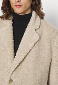 Persona con capelli ondulati fino alle spalle, che indossa un cappotto di pile beige a trama su un maglione nero a collo alto.