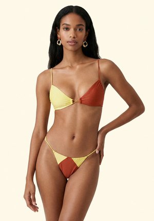 Donna che indossa un bikini a triangolo bicolore giallo e ruggine con sottili spalline, in piedi con una mano sul fianco davanti a uno sfondo semplice.