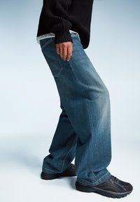 Svetlo modri jeans z ohlapnim krojem, ki imajo klasičen pet-žepni dizajn in subtilne detajle šivanja, v kombinaciji s temnimi športnimi čevlji.