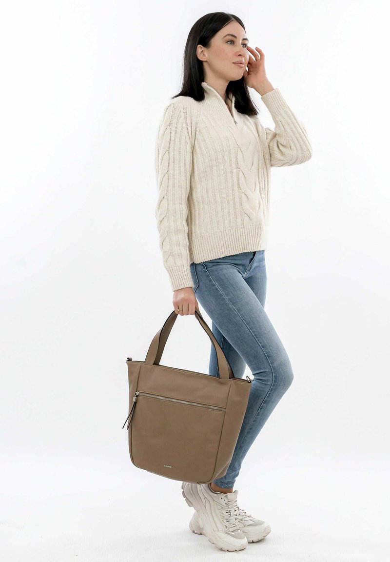 Bruine totebag met dubbele handvatten, gladde textuur, voorvak met rits, gecombineerd met een crèmekleurige kabelgebreide trui en lichtblauwe jeans.