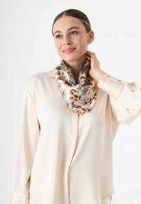 Écharpe en soie ornée d'un motif floral dans des teintes d'orange, de bleu et de rose, portée lâche autour du cou, sur une blouse beige.