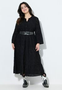 Musta maxi-mekko kukkakuosisella silkkireikäkankaalla, kolmanneksen pituiset hihat, napitettu etuosa, vyötäisillä kiristettävä helmassa olevat niitit ja kerroksellinen helma.