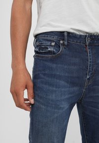 Man som bär mörkblå denimjeans och vit t-shirt, med handen avslappnad vid sin sida, midja och övre lår synliga.