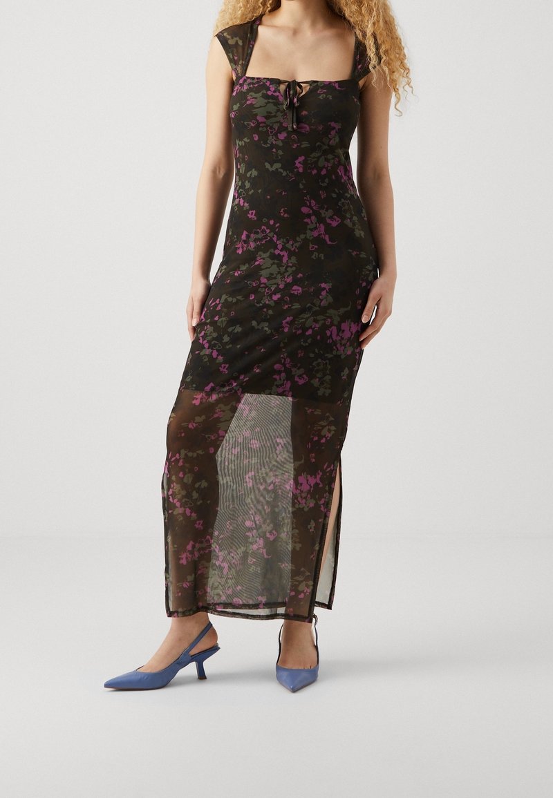 Schwarzes Blumenkleid mit kurzen Ärmeln, quadratischem Ausschnitt und Schnürdetail, mit einem transparenten Überwurf und seitlichen Schlitzen. Kombiniert mit blauen Absatzschuhen.