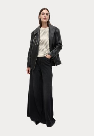 Chaqueta de moto de cuero negro sobre una camiseta de rayas beige y blanca, combinada con pantalones anchos negros de corte relajado. Estilo casual y cómodo.