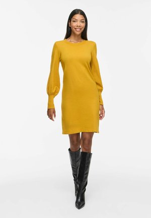 Femme marchant portant une robe jaune moutarde à manches longues avec des manches bouffantes et des bottes noires montantes jusqu'aux genoux sur fond blanc.