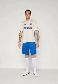 Muž stojí čelom dopredu a má na sebe biely a modrý futbalový dres FC Porto s logom Betano, biele ponožky a kopačky na pozadí bez vzoru.