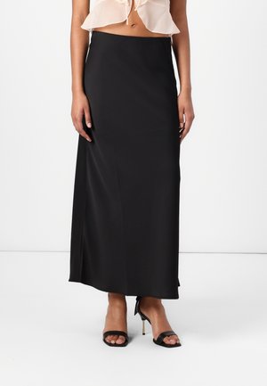 Gonna maxi nera in satin con una texture liscia, dotata di vita aderente e una leggera apertura laterale. Indossata con sandali neri con tacco alto e cinturini.